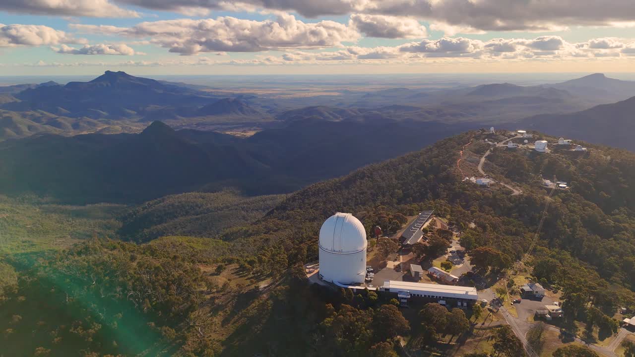 Drone footage glides above a mountaintop observatory, revealing telescopes, forested hills, and dramatic sunset lighting in Warrumbungle National Park, Australia