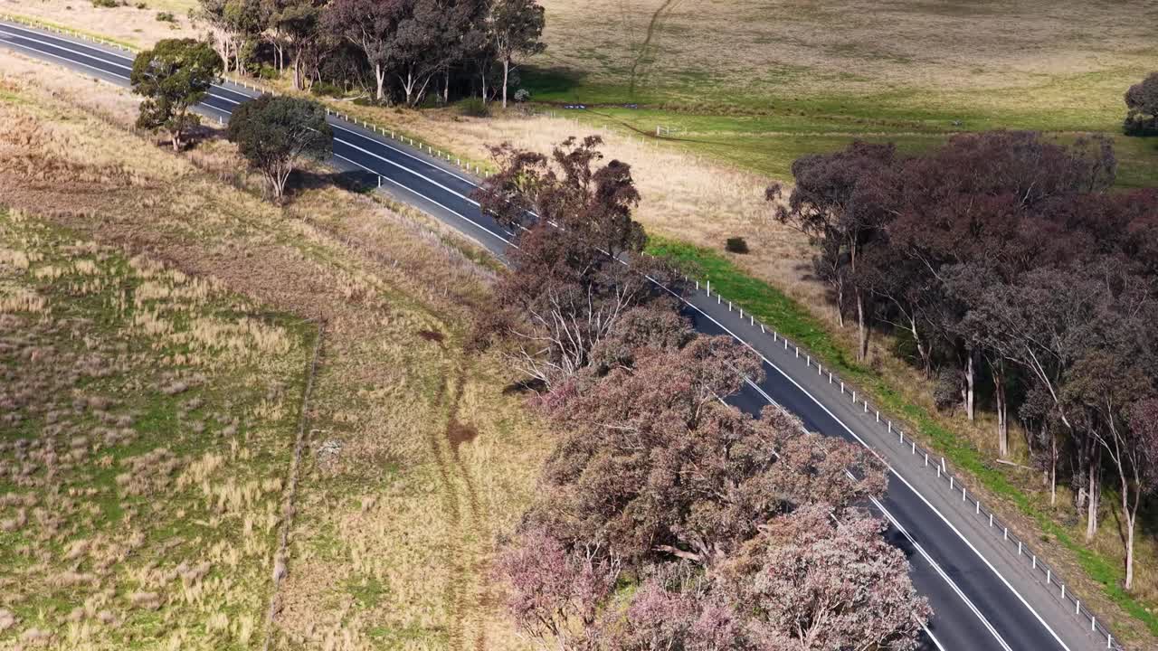 A single car travels along a winding country road bordered by trees and open fields, captured in daylight with smooth aerial camera movement and natural lighting