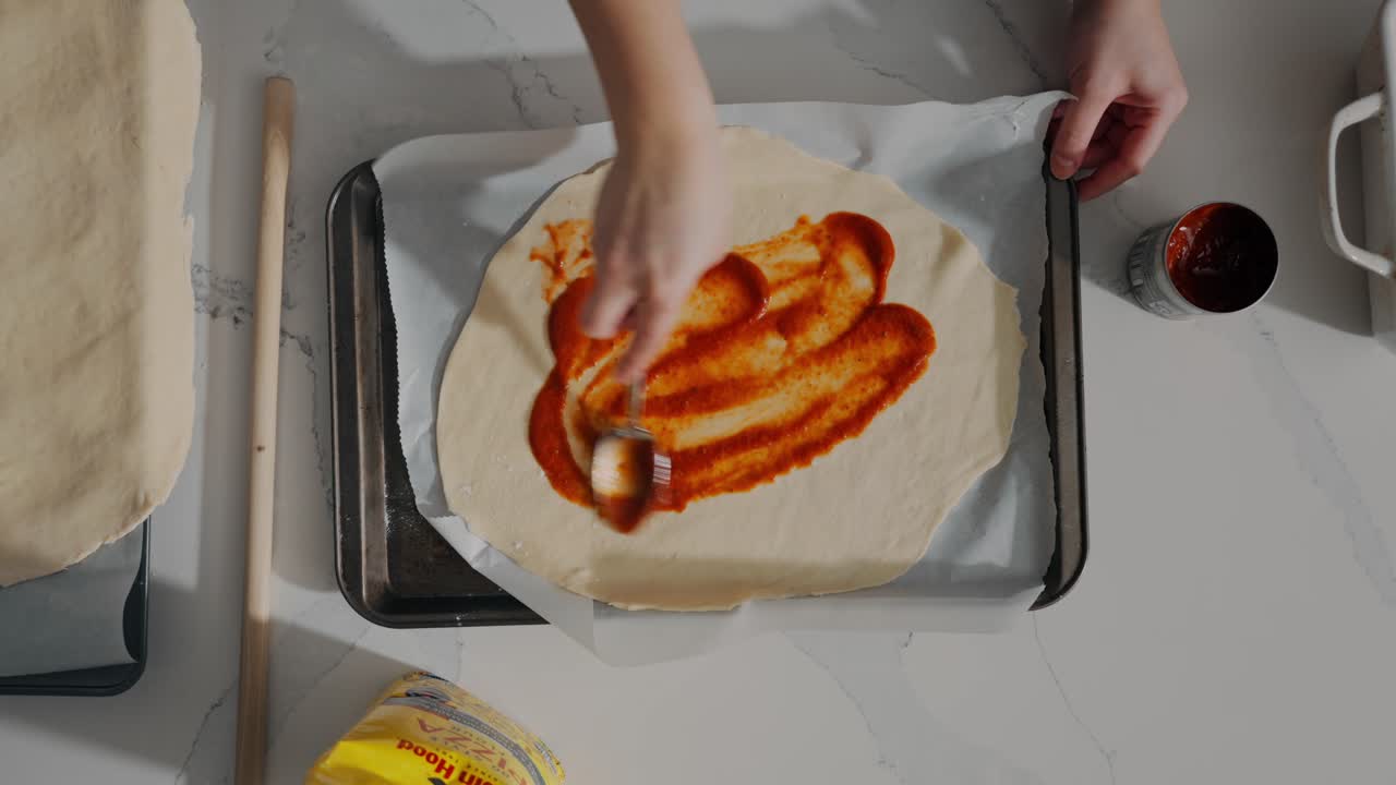 Oversiktsbilde av hender som sprer tomatsaus på pizzadeig før steking