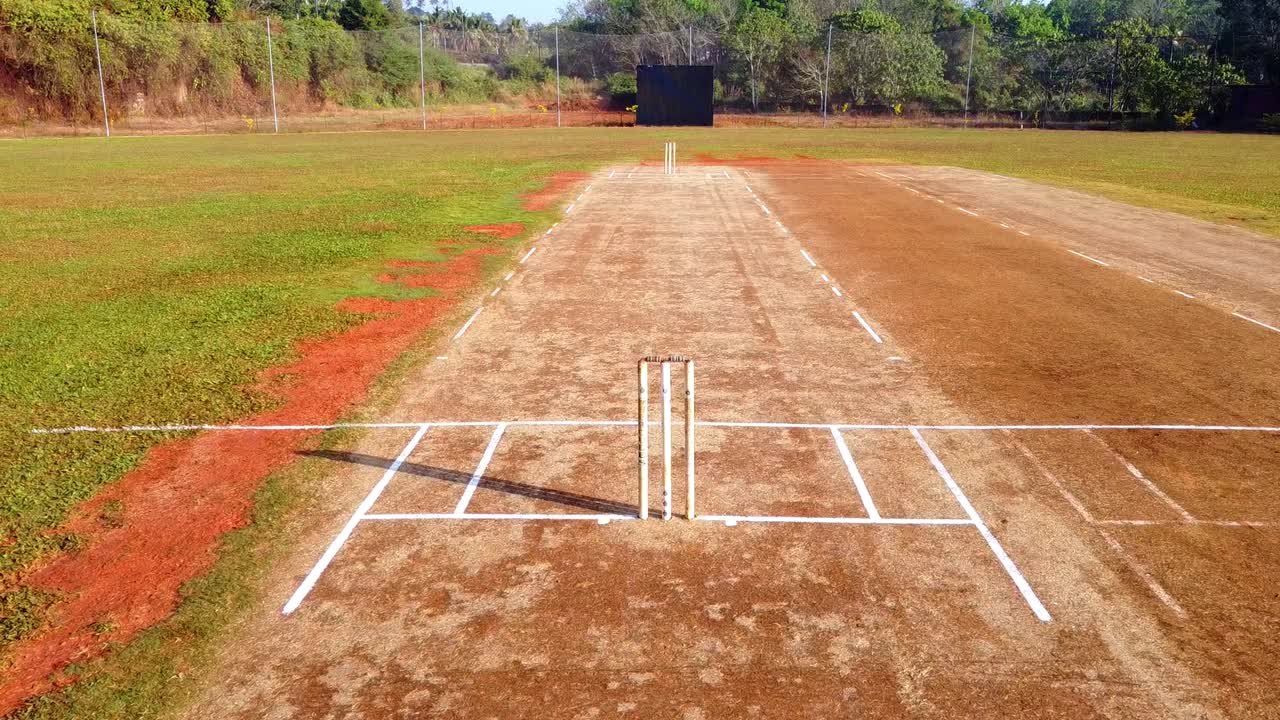 Drönarbild från ovan av en solig cricketplan, som zoomar ut från slagpinnarna för att visa hela planen och gränsen. Perfekt för teman inom sport och utomhusaktiviteter.