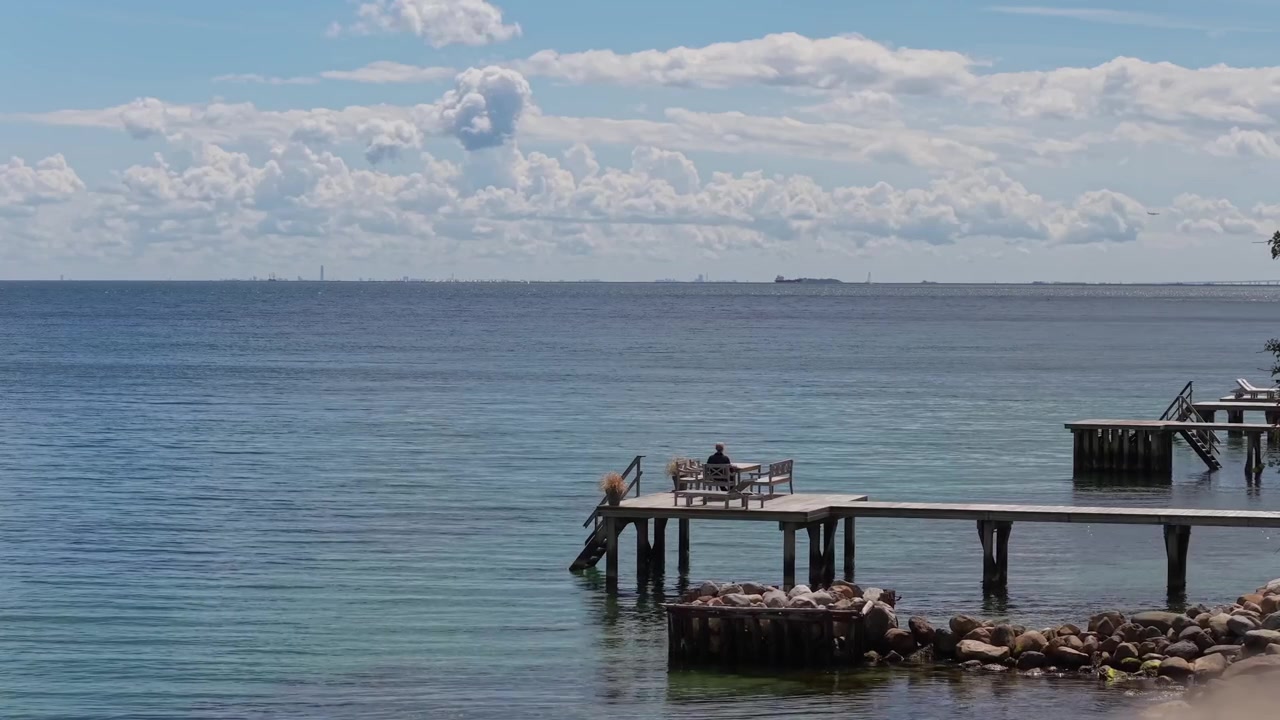 Peaceful coastal footage of an elderly man enjoying a quiet moment on the Taarbaek waterfront, overlooking the calm sea and distant horizon along Denmark's picturesque shoreline.