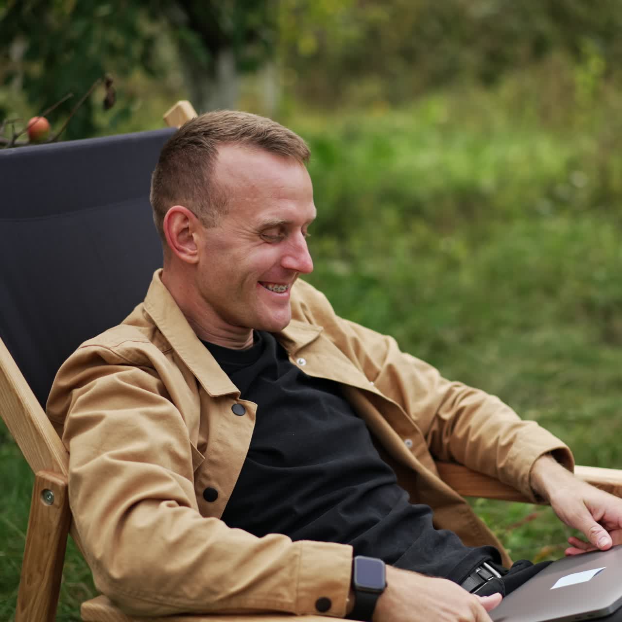 Happy smiling resilient man sitting in the chair outdoors. Male freelancer opens laptop and starts his remote online work. Nature backdrop