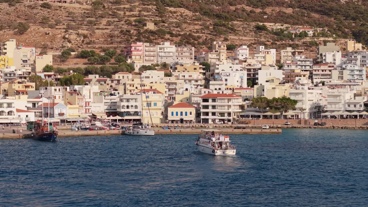 Cinematic drone footage captures a travel boat with tourists arriving at the scenic seafront of Karpathos, showcasing the island’s beautiful harbor and turquoise waters