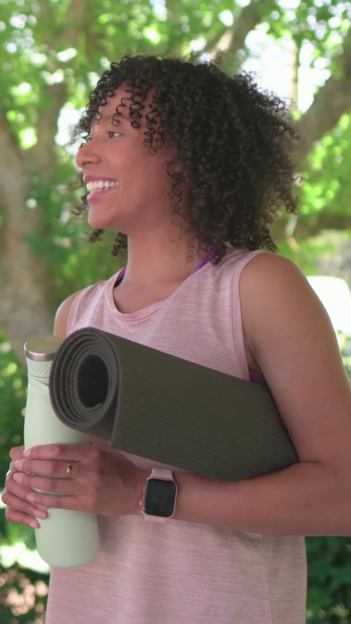 Vídeo vertical: Mulher de regata, segurando um tapete de yoga, reage ao parceiro enquanto abre uma garrafa de menta no jardim.