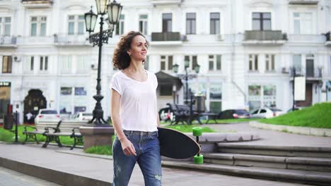 Smiling-attractive-girl-walking-in-the-old-city-street-holding-her-longboard-in-the-morning,-slow-motion-shot.-Beautiful-old