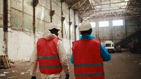 Rear-view-of-a-man-with-Black-skin-in-a-white-special-uniform-and-an-orange-vest-together-with-a-female-employee-walk-through-a-large-waste-processing-plant-and-point-with-his-hand-to-the-side