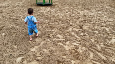 following the little kid in jeans romper walking barefoot by the sandy ground. lovely toddler heads to the merry-go-round.