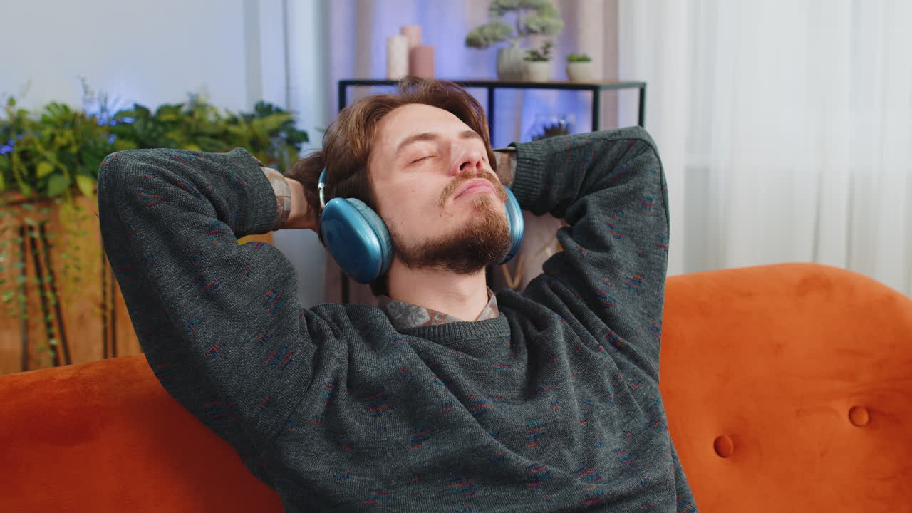 Uomo caucasico felice con cuffie wireless che balla ascoltando la sua musica rock 'n' roll preferita sul divano di casa
