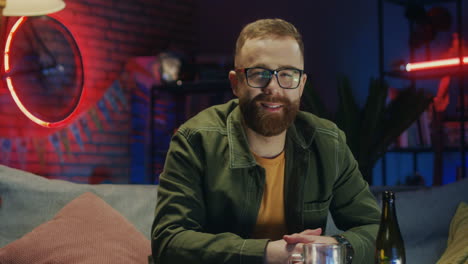 Portrait-Of-The-Joyful-Red-Haired-Young-Man-In-Glasses-Smiling-To-The-Camera-And-Being-In-Good-Mood-While-Sitting-On-The-Sofa-In-The-Dark-Room