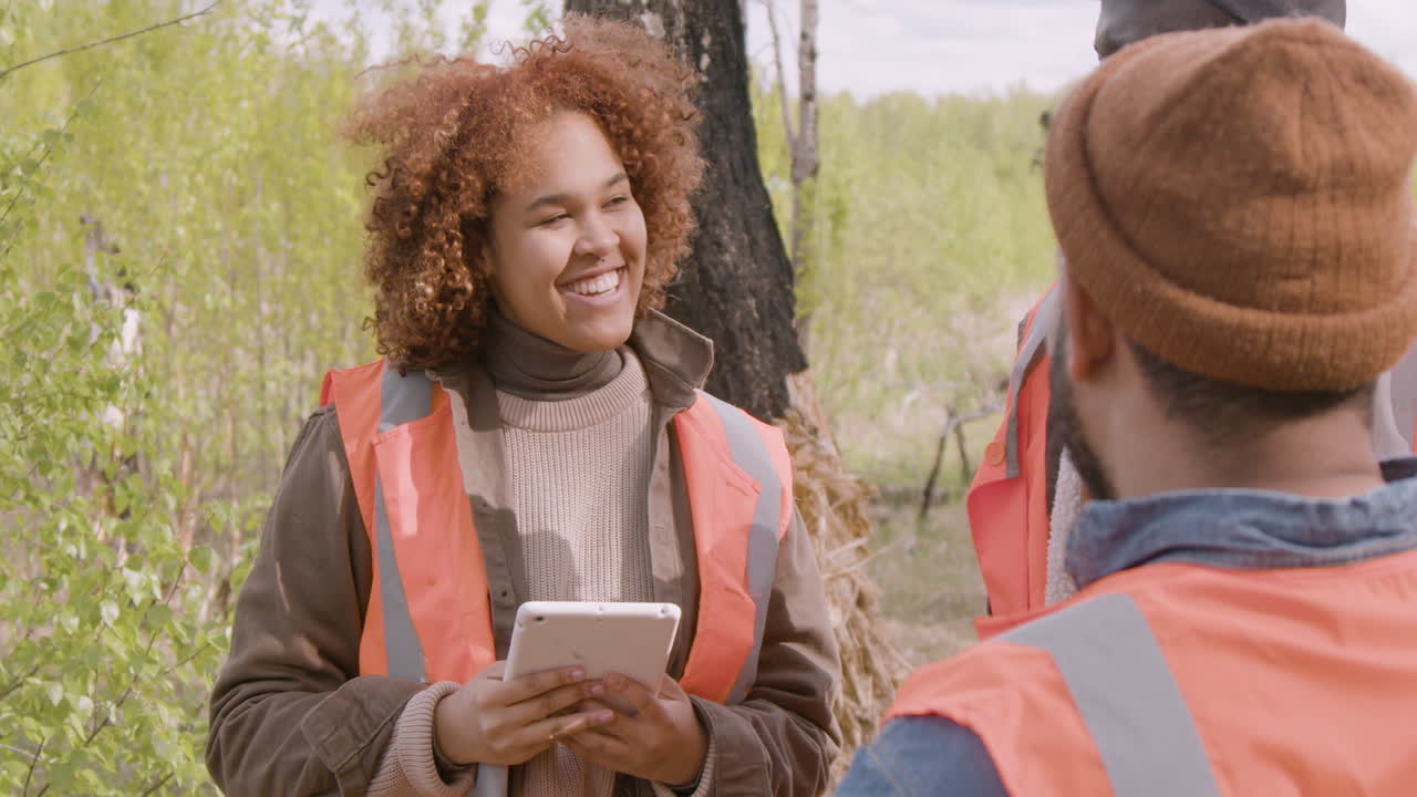 vista de cerca de una activista afroamericana sosteniendo una tableta y hablando con sus compañeros de trabajo en el bosque mientras deciden dónde plantar árboles