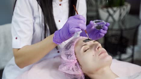 A beautician applies a black mask on the face of the client's face in a beauty salon. Skin care. Beauty industry