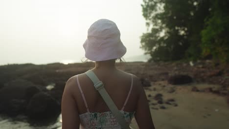 Young-woman-wearing-a-hat,-seen-from-behind,-facing-the-beach-at-sunset