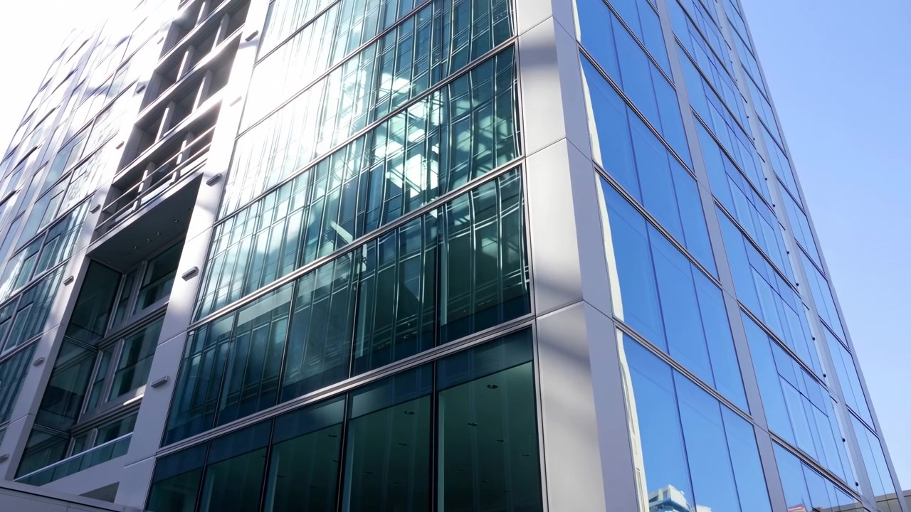 Vista em movimento de baixo ângulo de um canto de torre de escritórios contemporânea com fachada de vidro e estrutura metálica, enquanto a luz do sol desliza pelas superfícies, criando reflexos brilhantes, forte contraste e geometria urbana limpa
