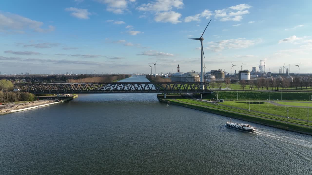Zoom in aerial shot of Antwerp industrial canal with bridge crossing, boat navigating, and wind turbines in the background on sunny day