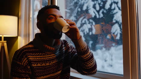 man drinking a warm beverage by a window during a snowy winter day