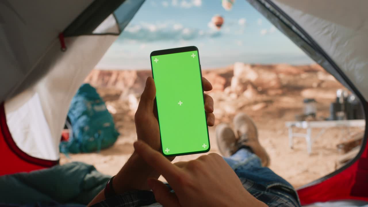 vista de un turista sosteniendo verticalmente un teléfono inteligente con un marcador de posición de pantalla verde, deslizando un dedo sobre la pantalla. viajero descansando en una tienda en la cima de una montaña rocosa y volando globos de aire caliente.