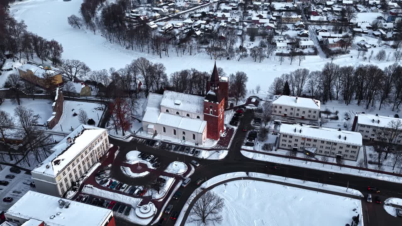 Chiesa storica e città invernale con edifici innevati e fiume ghiacciato, in vista aerea.
