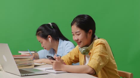 close up of asian woman student enjoys using smartphone while sitting with her friend studying on a table with laptop in the green screen background classroom