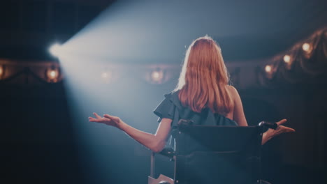 young actress on wheelchair speaking to audience pan left view of young woman smiling and gesticulating while sitting on wheelchair in spotlight and talking to audience during performance in dark theater