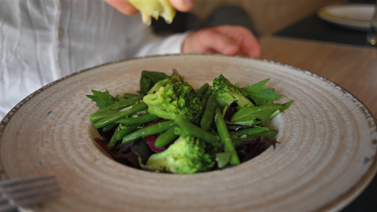 Hand squeezing fresh lemon over green vegetable salad with broccoli and beans