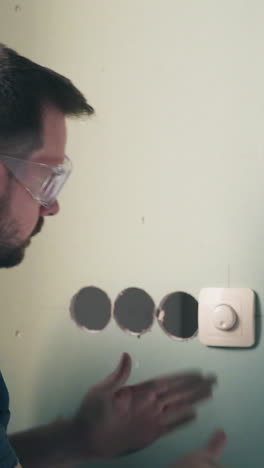 professional electrician in blue shirt installs modern lever in white plastic case on green wall and poses to camera in flat