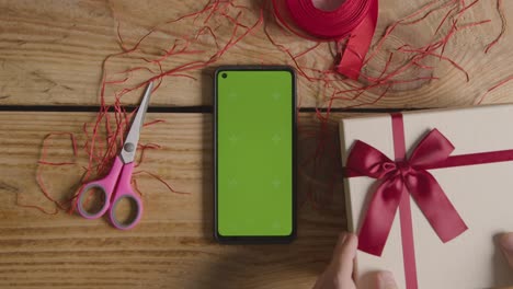 Overhead-Shot-Of-Man-Gift-Wrapping-Romantic-Valentines-Present-Of-Perfume-In-Box-Next-To-Green-Screen-Mobile-Phone