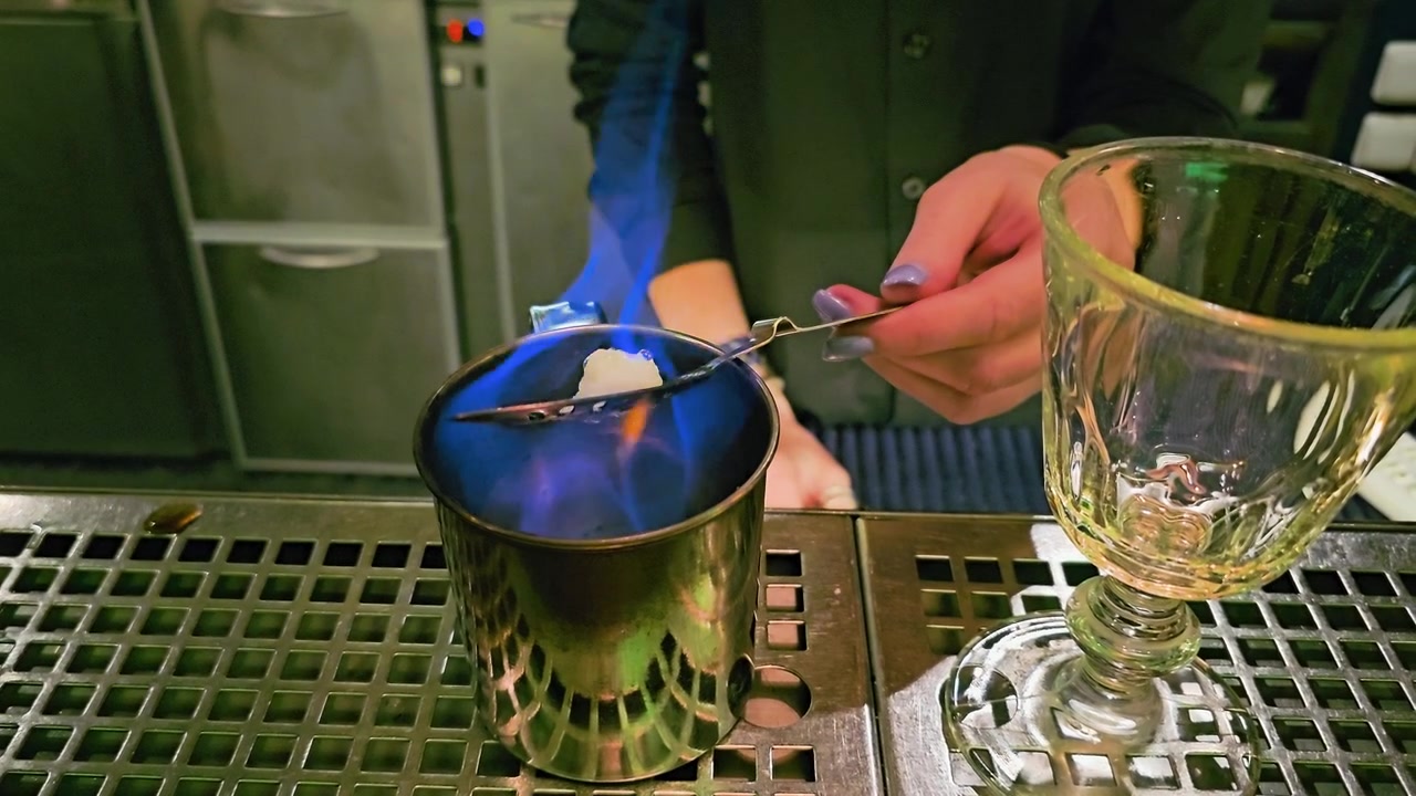 Primo piano di un cocktail lounge buio che mostra un rituale dell'assenzio, fiamme blu, una zolletta di zucchero, un cucchiaio forato, un bicchiere miscelatore in metallo, un calice vuoto e le mani del barista sopra una ringhiera del bar in acciaio inossidabile.