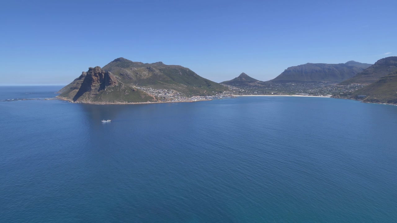 Et malerisk skud fra Chapman’s Peak Drive langs Cape Peninsula, der afslører en fisketrawler, der forlader Hout Bay havn, med dramatiske bjerge og betagende kystlandskab.