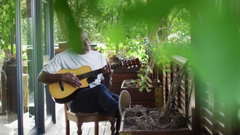 Thoughtful-african-american-senior-man-sitting-on-balcony-playing-acoustic-guitar