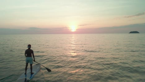 drone rastreando a un hombre de pie remando al atardecer en el océano con una isla en la distancia