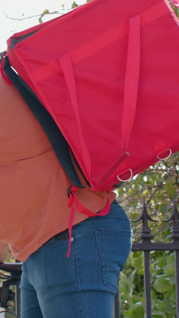 Video verticale: Un uomo vede una borsa rossa, la afferra, la solleva e se la mette sulla schiena su un sentiero per la consegna in bicicletta.