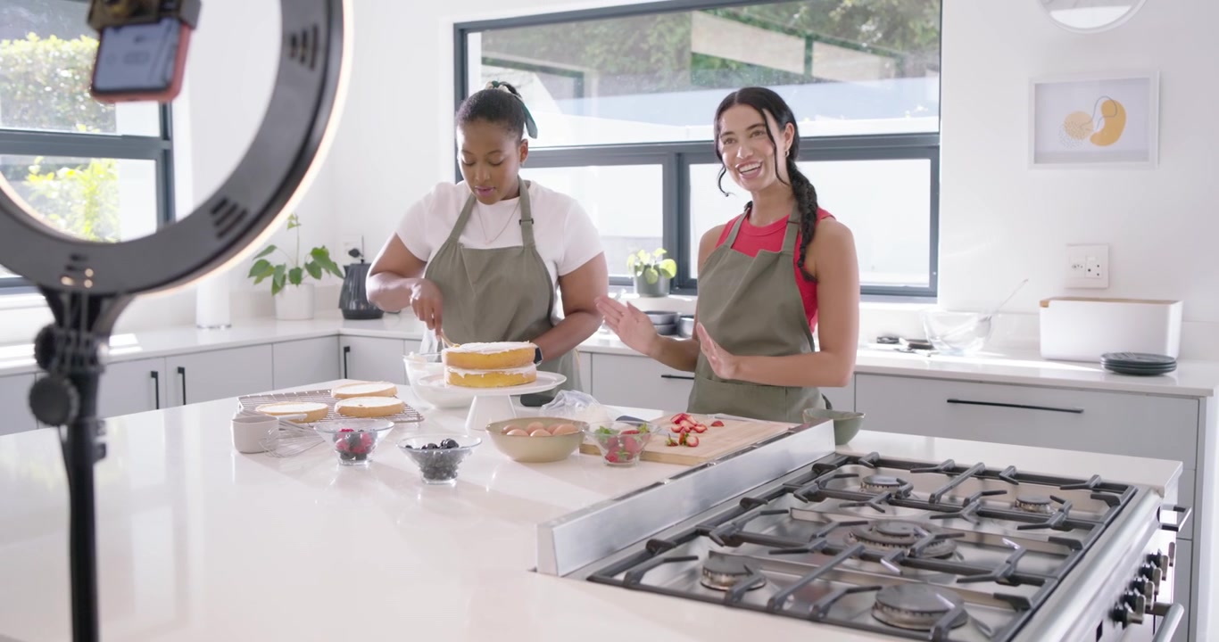 Apresentadoras diversas de avental confeitando bolo em uma ilha de cozinha enquanto um operador enquadra a filmagem com DSLR e ring light.