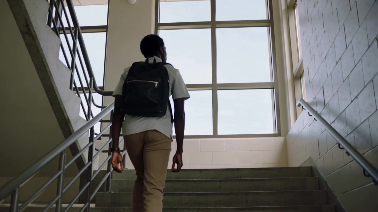 En elev i hvid skjorte og khakis starter opstigningen ad skoletrappen med en mørk rygsæk mod vinduet.