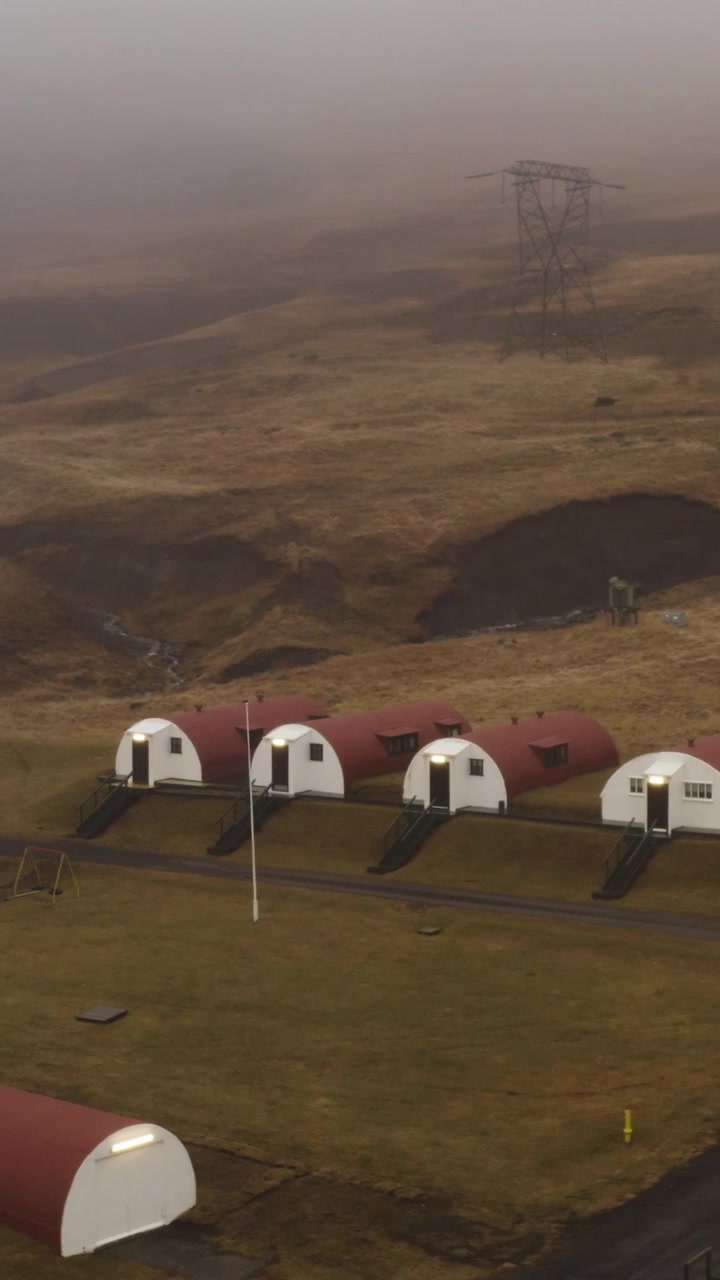 Luftfoto av unike rødtakede Nissen-hytte-overnattingssteder i en avsidesliggende, gresskledd dal i Hvalfjörður, Island. En stor strømmast står på en tåkete ås i bakgrunnen