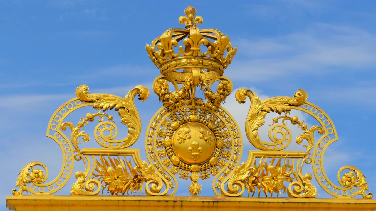 Ornate golden crown and fleur-de-lis symbols adorning the royal gate at the palace of versailles under a blue sky.