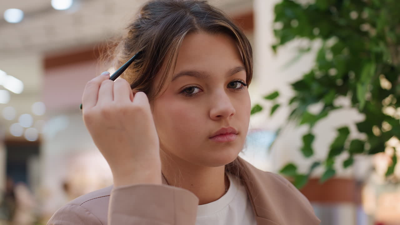 Una joven sentada en un centro comercial cerca de una planta, ajustándose el pelo y mirando pensativa las cálidas luces bokeh de las fachadas de las tiendas al fondo, con una chaqueta beige y una camiseta blanca, ambiente cinematográfico íntimo.
