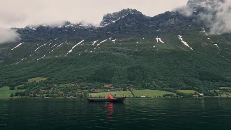 Person-Mit-Roter-Jacke,-Rudern-In-Einem-Kleinen-Boot-Auf-Einem-See,-Große-Berge-Und-Wolken-Im-Hintergrund