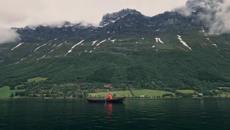 persona con chaqueta roja, remando en un pequeño bote en un lago, grandes montañas y nubes en el fondo