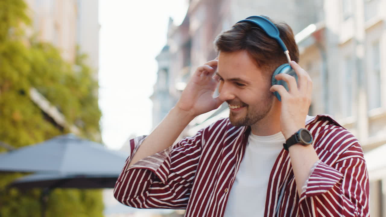Happy bearded man in wireless headphones choosing listening music dancing outdoors city street