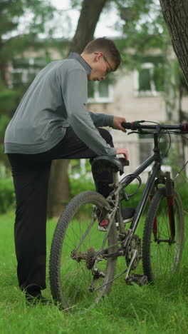 un niño camina a una bicicleta estacionada cerca de un árbol en un campo cubierto de hierba, revisa la bicicleta, toca el pedal con su zapato y coloca su pie en el pedal mientras se centra en algo en su pie