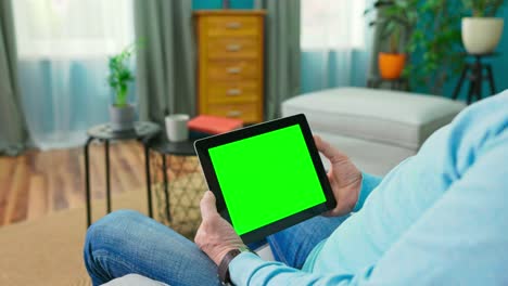 primer plano de una tableta digital con una pantalla de simulacro verde en blanco sostenida por un anciano adulto relajándose en una habitación acogedora en casa. la cámara se aleja de la tableta