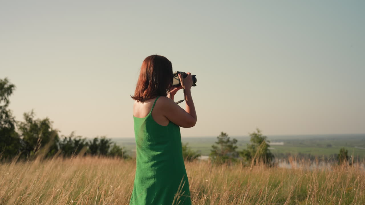 woman photographer in green dress walks through tall grass holding camera as she lifts view to distant horizon under clear sky during calm golden hour in countryside