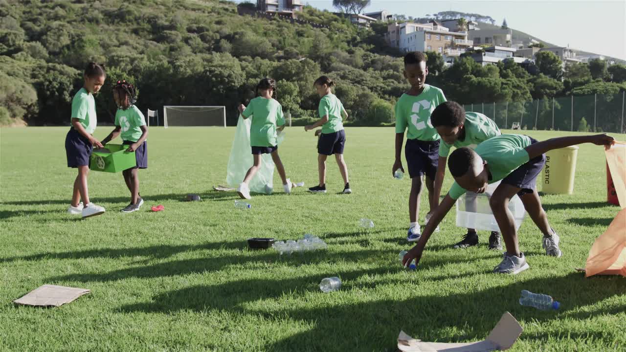 재활용 티셔츠를 입은 다양한 행복한 학교 학생들이 초등학교에서 스포츠장을 청소하고 있습니다.