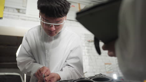 close up of student in lab coat wearing protective face shield, using vernier caliper to measure engine component in automotive workshop, with partial view of colleague recording data on tablet