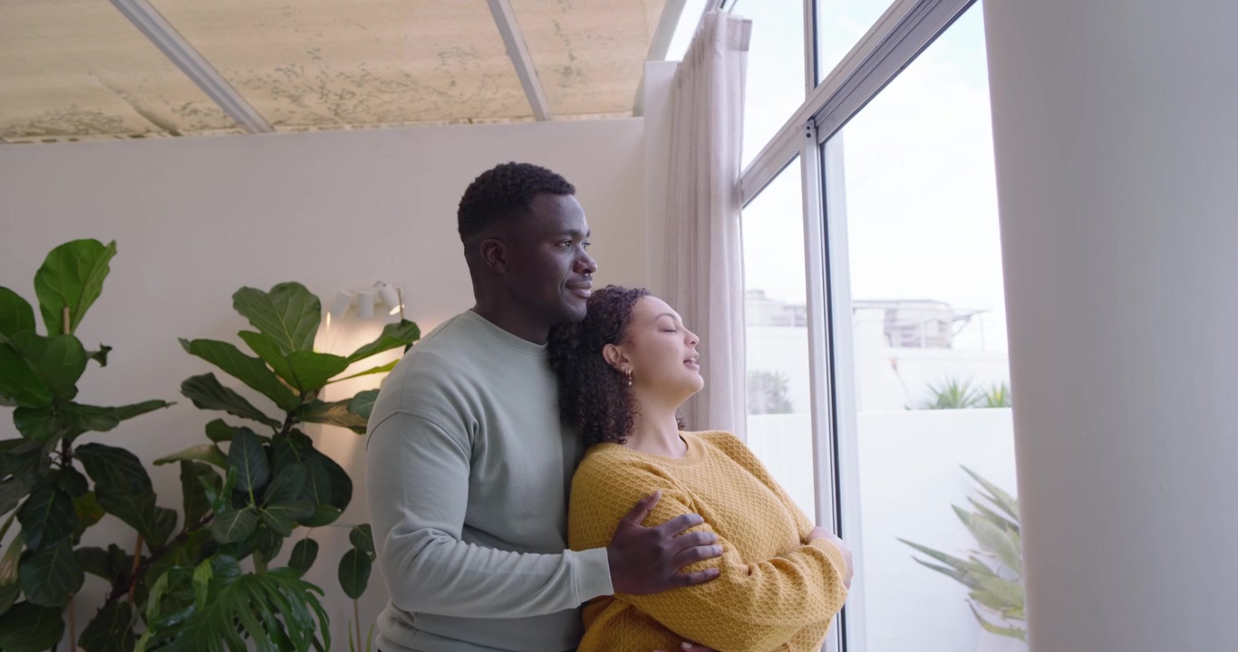 Couple afro-américain près de la fenêtre en pulls, l'homme étreignant la femme qui se penche en arrière en souriant et partageant la vue