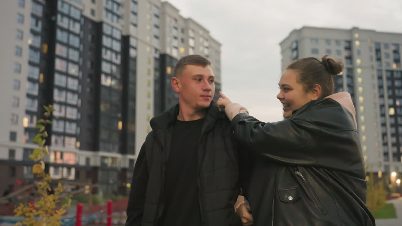 Siblings enjoy fun moment walking together as sister gently tickles brother to make him laugh while both smile warmly surrounded by tall buildings and soft city lights during cool evening stroll