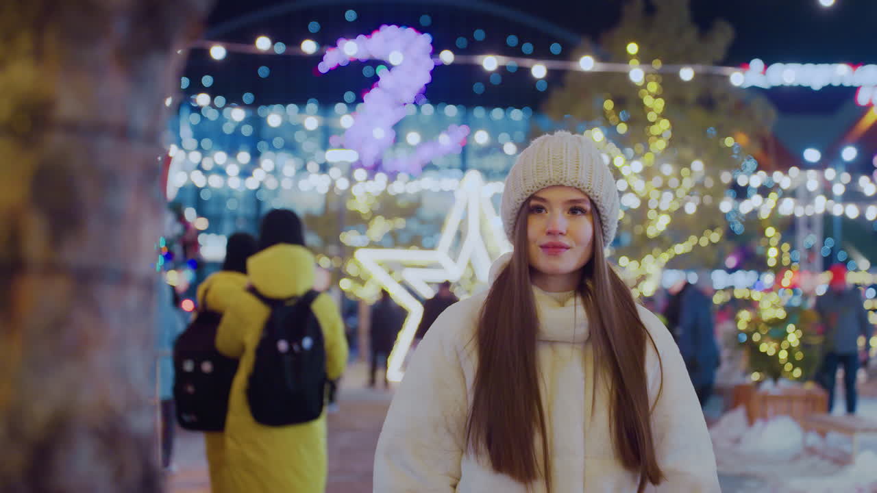 Smiling woman in cozy winter outfit admiring beautifully lit tree and star decorations at night with people walking around in a festive setting