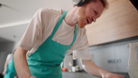 a confident blond guy in a white t-shirt and a blue apron washes the table in the kitchen and dances while listening to music on wireless headphones during his on-call cleaning in a modern apartment