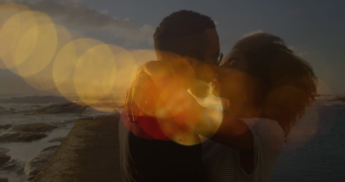 Animation of lens flares over diverse couple kissing and hugging on beach against sea and sky