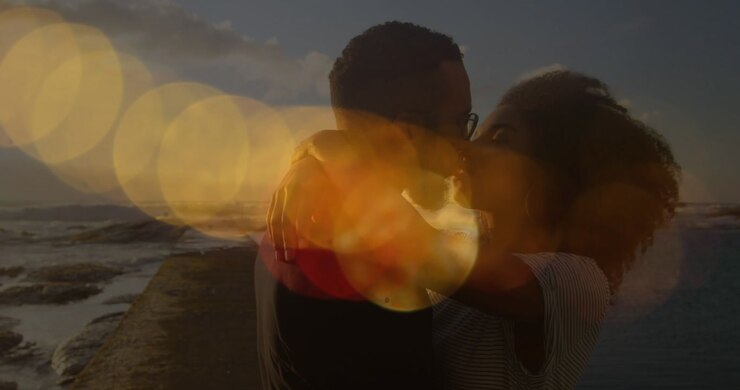 Animation of lens flares over diverse couple kissing and hugging on beach against sea and sky
